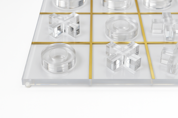 Close-up of lucite Tic Tac Toe game with gold grid and clear pieces on white background